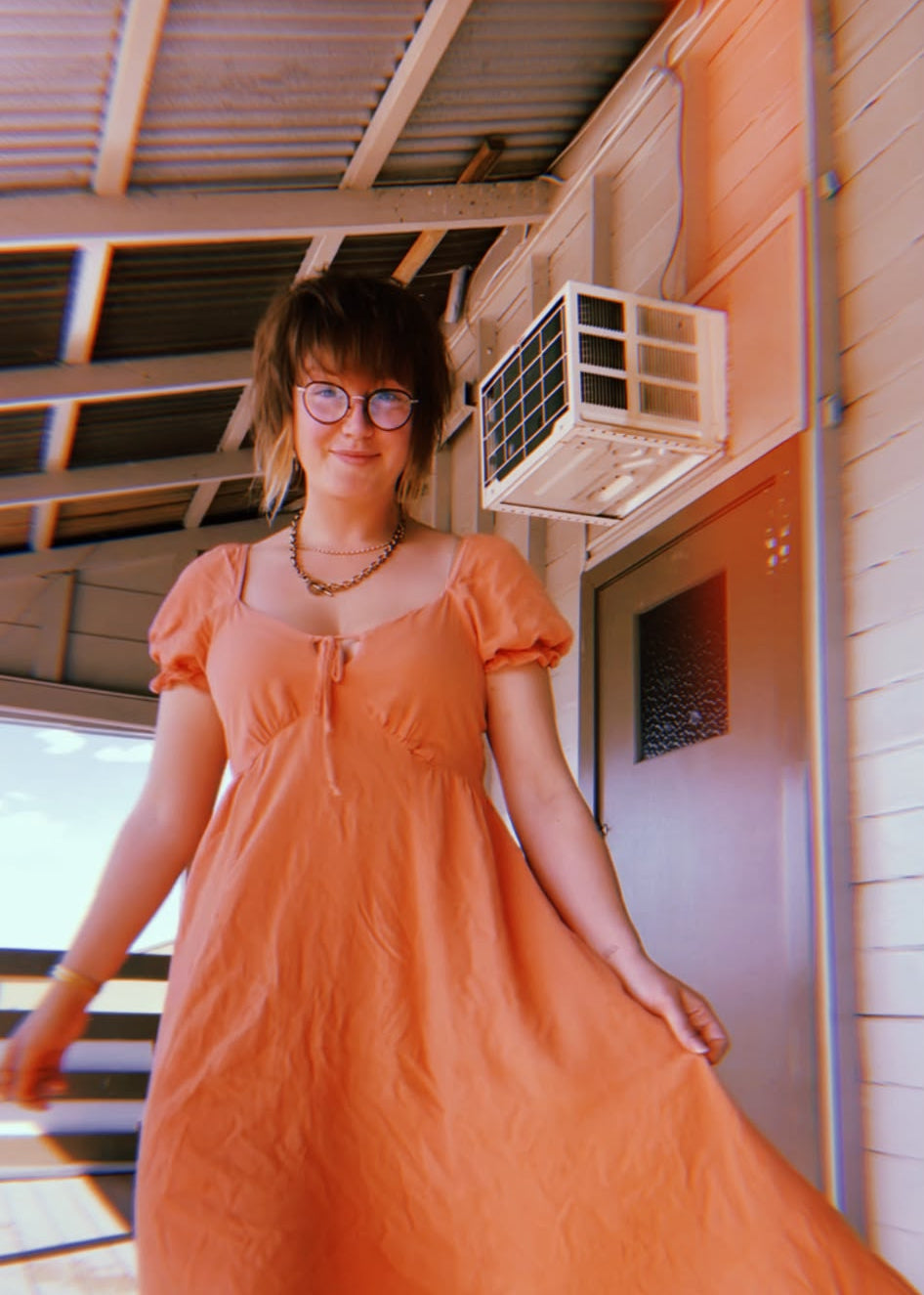 Bay Jemima is wearing a light orange puff-sleeve midi dress with a ruffled hem, standing on a wooden deck under a house eaves with an air conditioner visible in the background.