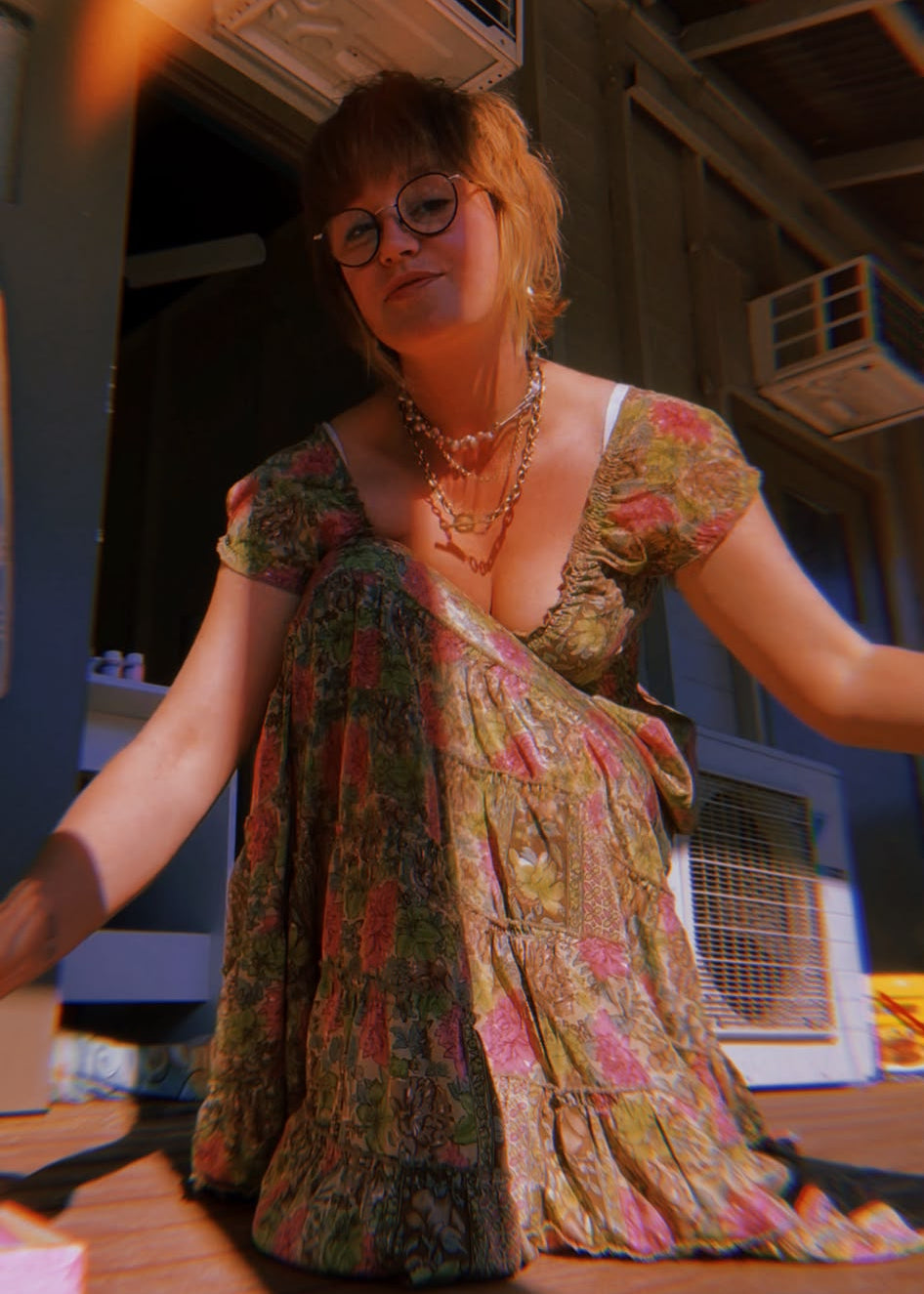 A woman with glasses and short auburn hair sits on a wooden floor wearing a bohemian floral maxi dress with short sleeves and a deep V-neck. The dress features a green, pink, and yellow pattern.